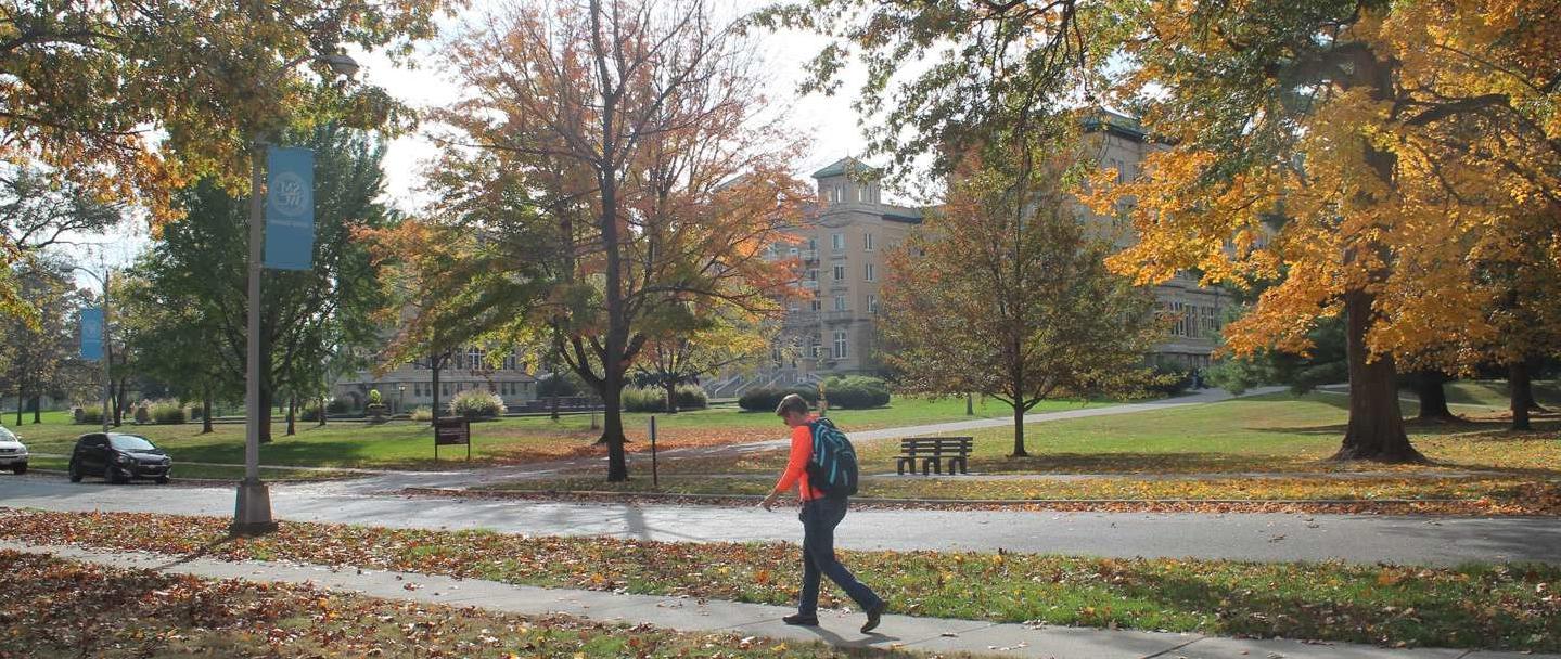A student walks across campus with the fall leaves and Le Fer Hall in the background.